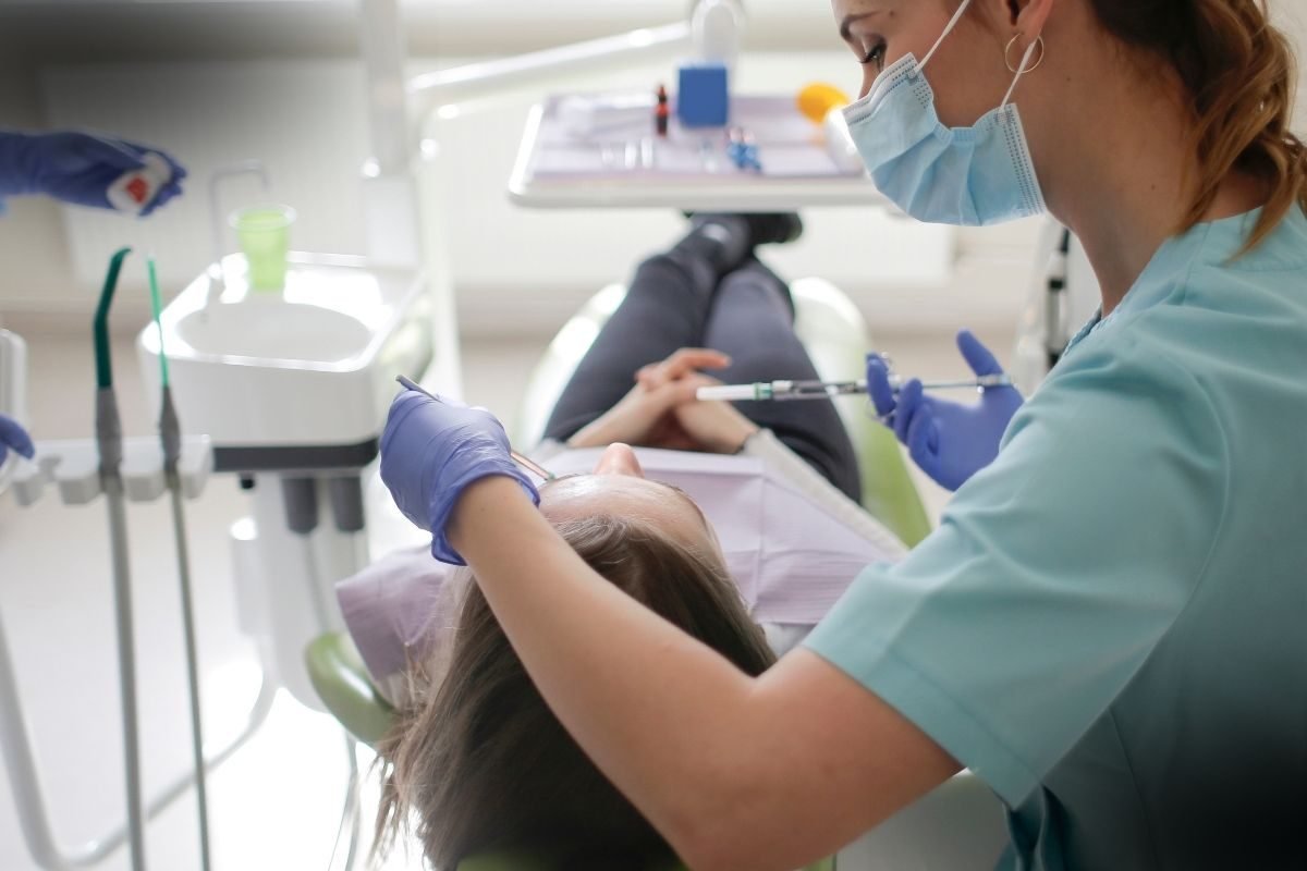 Indenização por procedimento odontológico: Dentista de luvas azuis realizando tratamento em paciente deitado na cadeira odontológica, com equipamentos médicos ao fundo.