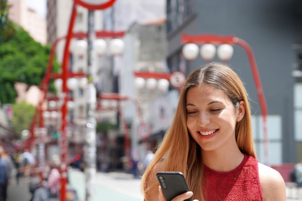 Consulta Advogado de Família em São Paulo (SP): uma mulher sorridente usando um vestido vermelho consulta seu smartphone, com a característica arquitetura urbana e a vibrante decoração de rua do bairro da Liberdade ao fundo desfocado.