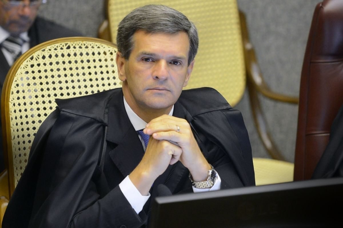 Mudança no Registro: Ministro de toga preta e gravata azul sentado, com expressão séria, olhando para frente, mãos entrelaçadas na frente do corpo em uma sala de audiência.