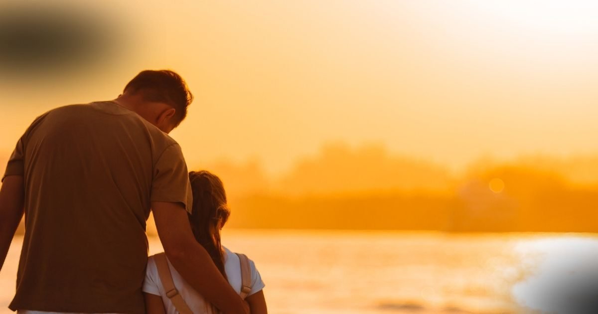 Pai conquista guarda da filha, mostrado por um pai de camiseta bege consolando sua filha ao pôr-do-sol, com o céu alaranjado ao fundo